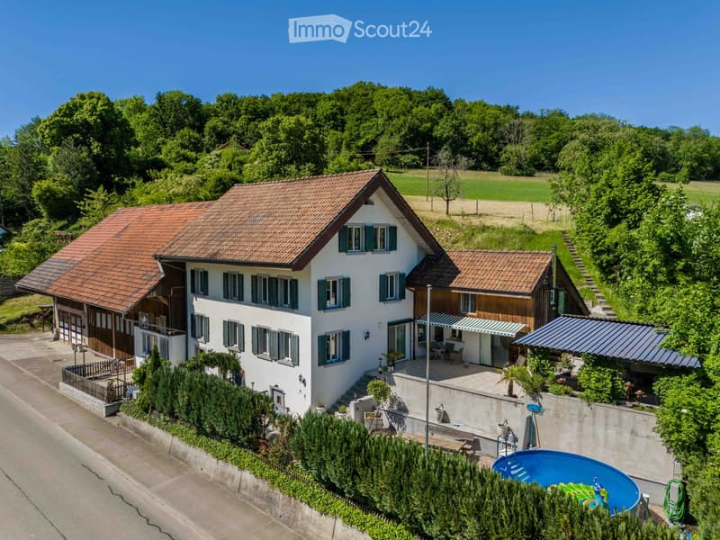 Two-story house with red-tiled roof, white walls, green shutters, and a garage. The property includes a swimming pool, patio, and garden area with trees and shrubs.