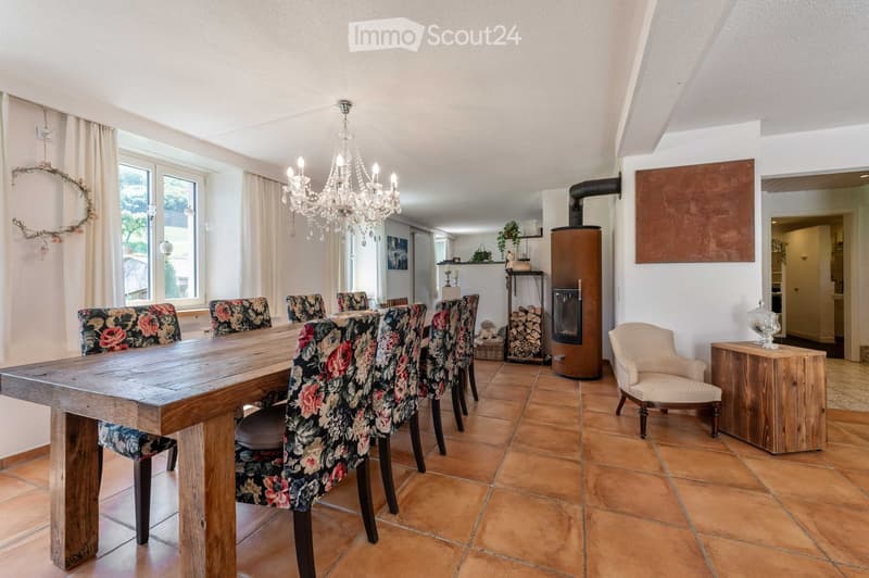 Dining room with a long wooden dining table surrounded by floral-patterned chairs, a chandelier hanging from the ceiling, a wood stove, and a small side table with a decorative item.