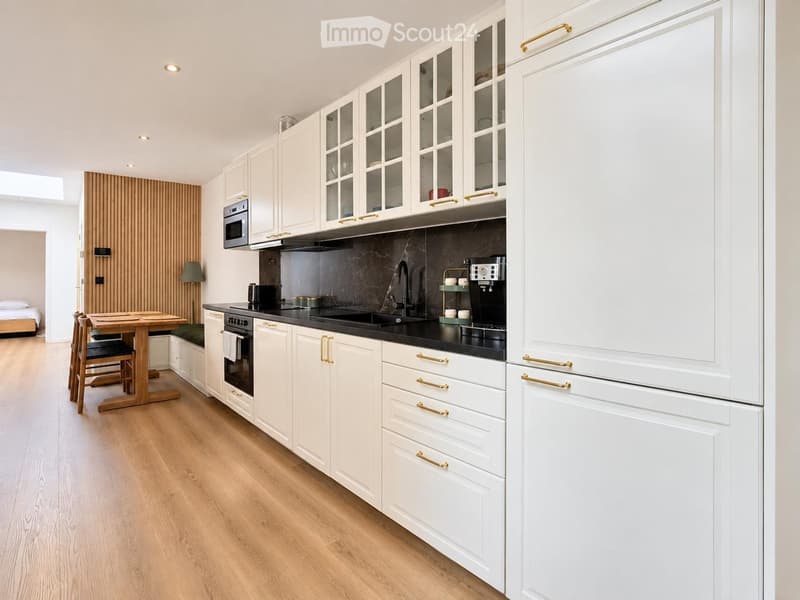 Modern kitchen with white cabinets, black countertops, and stainless steel appliances. Includes a wooden dining table with chairs, a coffee maker, and a microwave.