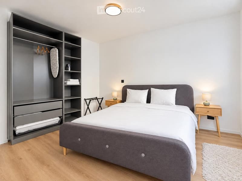 Modern bedroom with a large bed, grey upholstered headboard, white bedding, and two nightstands with lamps. A tall, grey wardrobe with shelves and drawers is on the left, and a small wooden stool is near the bed.