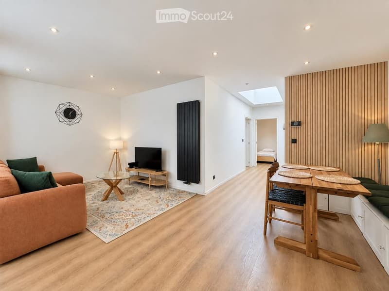 Modern living room with a sofa, coffee table, dining table, chairs, TV, and decorative lamp. The space features light wooden flooring, white walls, and a skylight above.