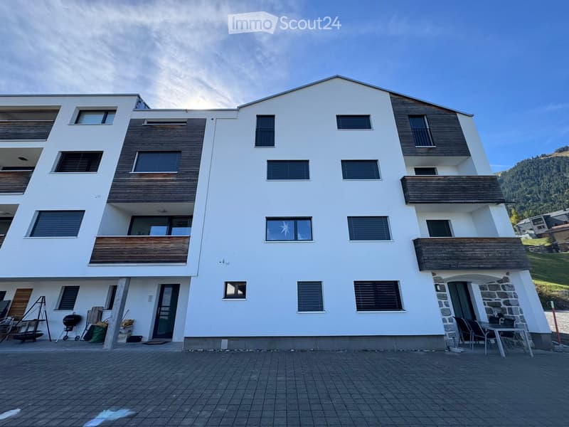 Modern multi-story building with white walls, wooden accents, and multiple balconies. Features include large windows, a stone base, and outdoor seating area.