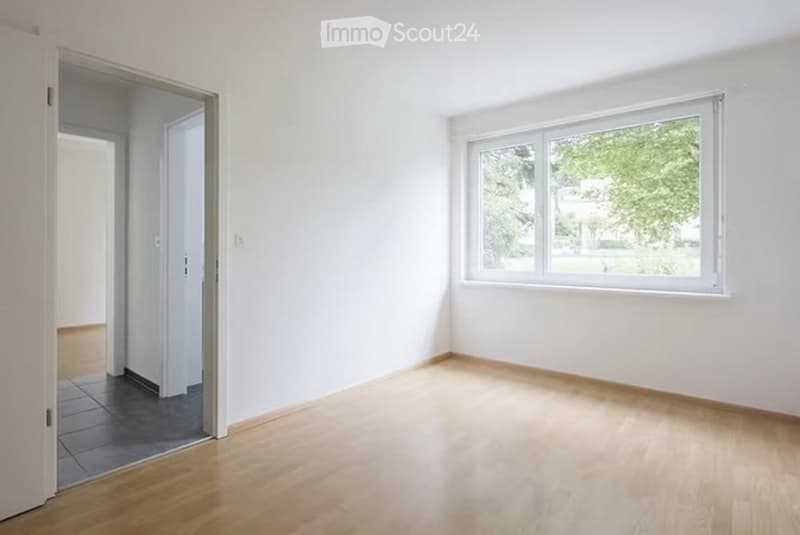 Empty room with white walls, light wooden flooring, and a large window with a view of greenery outside. The room has an open doorway leading to another space.