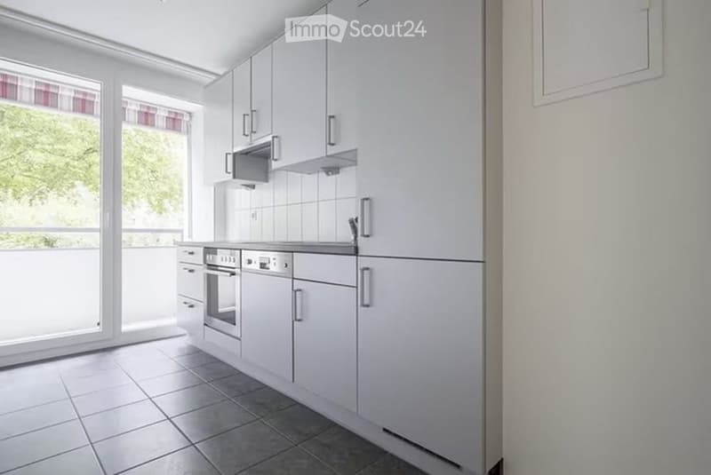Modern kitchen with white cabinets, stainless steel appliances, tiled floor, and large windows offering a view of greenery.
