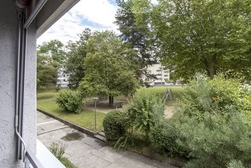 View from a balcony or terrace showing a well-maintained garden area with trees, shrubs, a small playground, and a paved walkway.