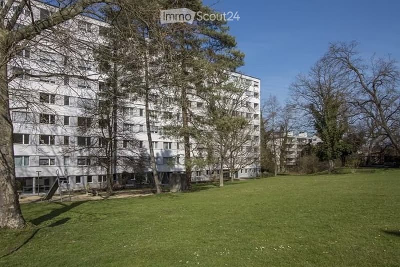 A large, multi-story residential building with numerous windows, surrounded by trees and a grassy lawn.
