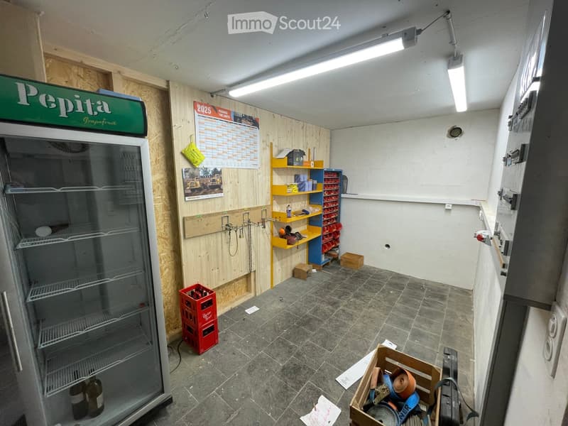 A small, partially furnished room with a refrigerator, shelves holding various items, storage boxes, and a calendar on the wall.