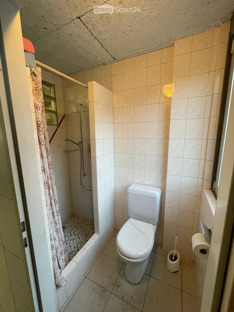 Small bathroom with a toilet, shower, and toilet paper holder. The shower has a curtain, and the walls are tiled.