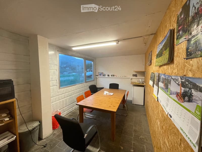 Office space with a wooden table, chairs, a screen, and several calendars on the wall. The room has large windows with a view of the outside and a small shelf with various items.