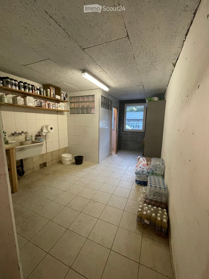 A narrow hallway with tiled floors and walls, featuring shelves with cleaning supplies, a sink, and a window at the end.