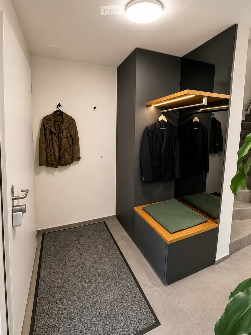 Modern hallway with built-in dark grey storage unit, wooden bench with green cushions, coat rack with jackets, and a grey rug on the floor.