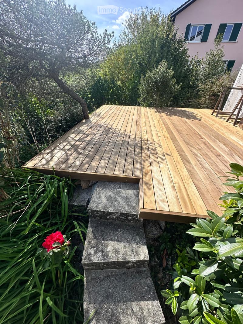 Wooden deck with stone steps leading up, surrounded by greenery and a red flower, with a house visible in the background.