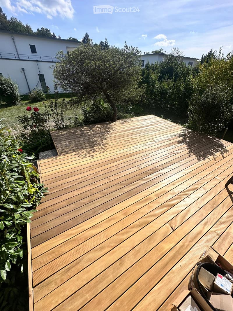 Wooden deck area with surrounding greenery, including bushes and trees, with a modern white building in the background.