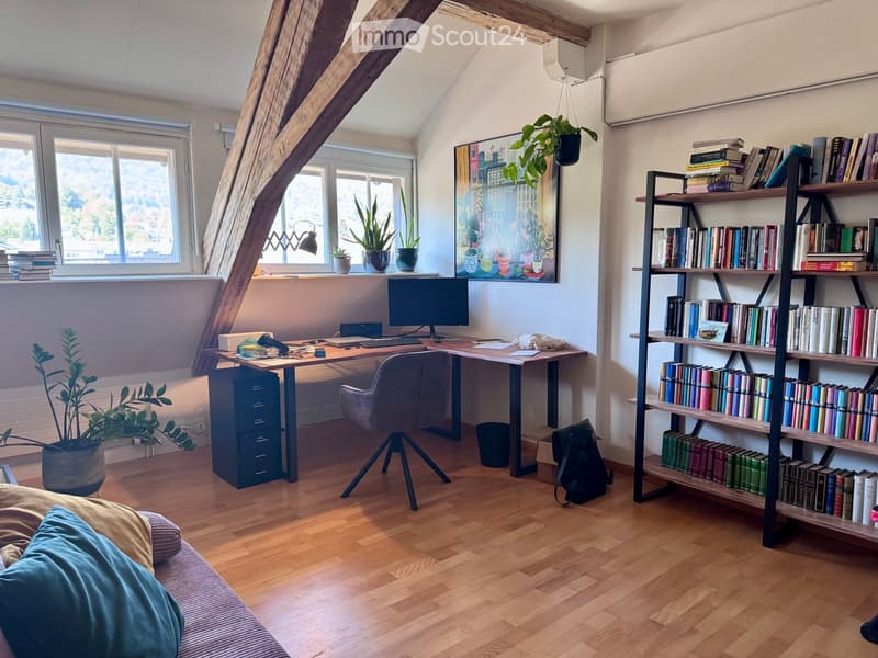 A cozy, multi-functional room with a slanted wooden ceiling, featuring a sofa, a desk with a computer, a bookshelf filled with books, and potted plants. The room has large windows with a view and is well-lit.