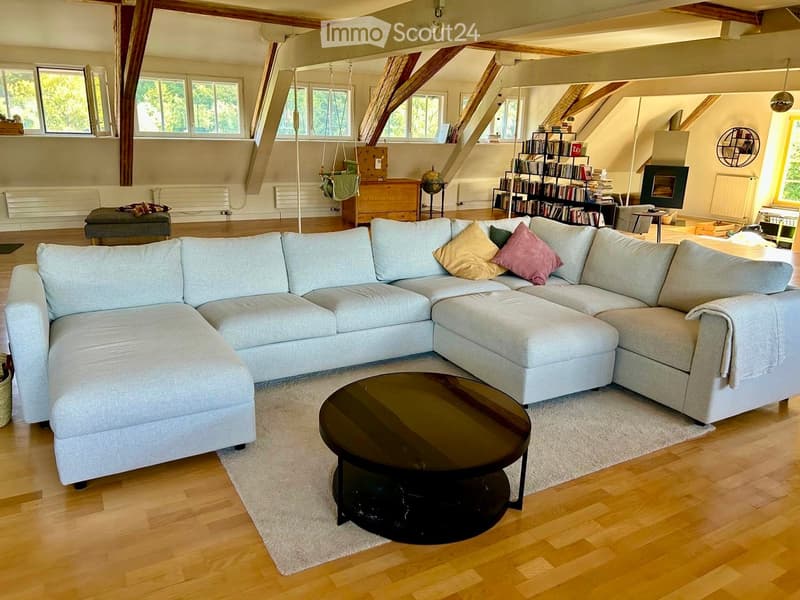 Bright living room with exposed wooden beams, large windows, grey sectional sofa, coffee table, bookshelf, and fireplace.