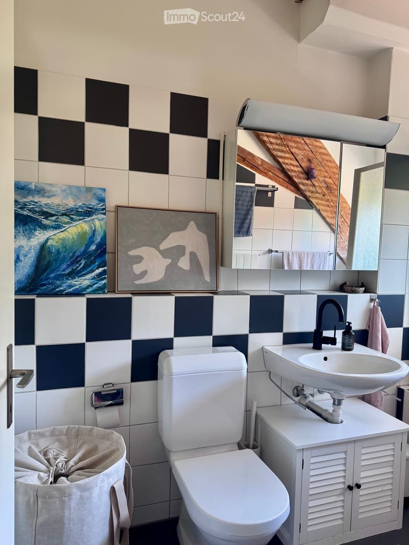 Modern bathroom with white tiles, a toilet, a sink with a modern faucet, a mirror cabinet, and decorative artwork on the wall.