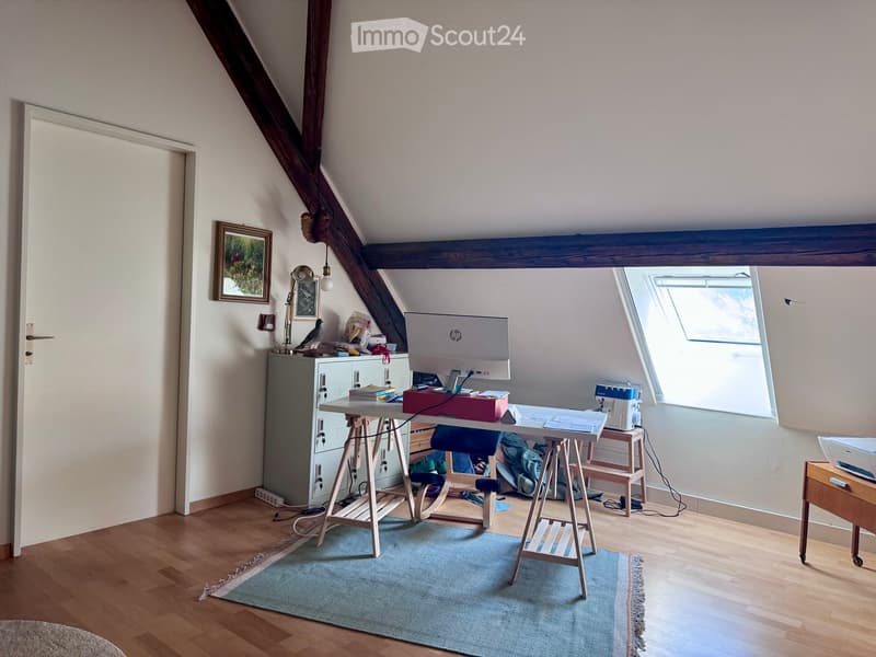 A well-lit attic room with wooden floors, slanted ceilings, and exposed wooden beams. It features a desk with a laptop, a chair, a lamp, a cabinet, a sewing machine on a small table, and a bird on a shelf. There is also a blue rug on the floor and a pictu