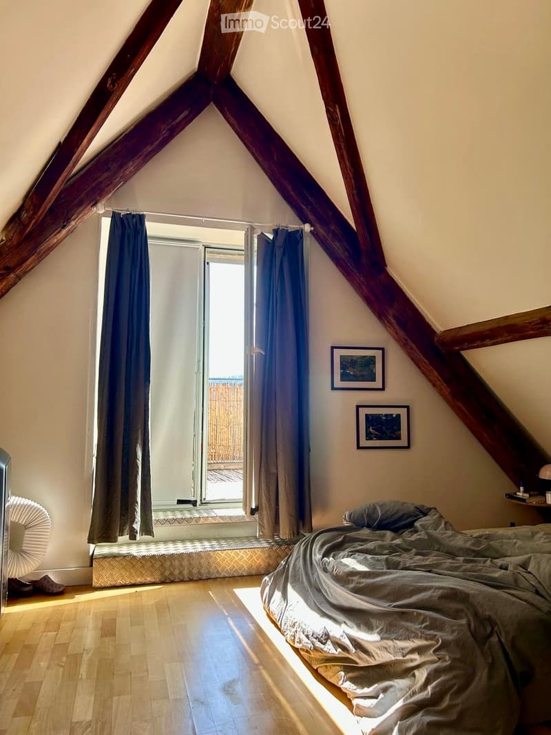 Cozy attic bedroom with wooden beams, a bed, a small seating area by the window, and a fan near the door.