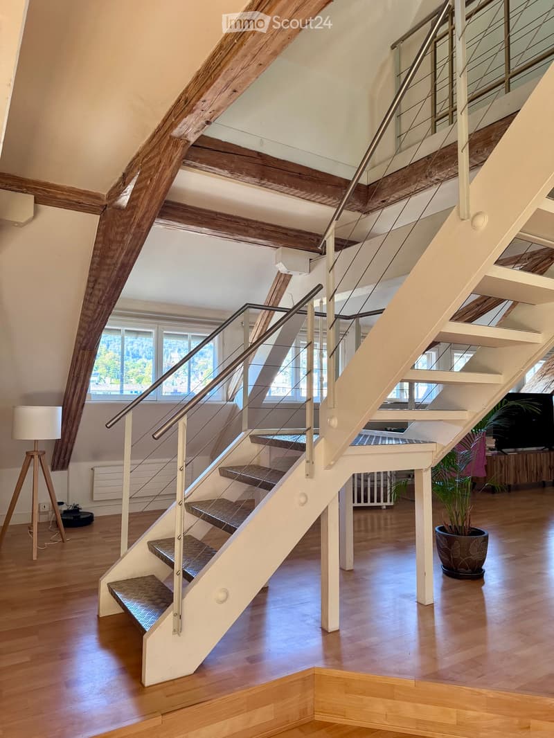 Spacious hallway with wooden floors, exposed wooden beams, and a modern staircase with glass railings. Large windows provide natural light, and there are potted plants and a lamp visible in the background.