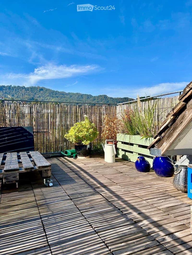 Wooden deck with a bench, potted plants, vases, and a bird figurine, surrounded by a wooden fence and a scenic mountain view.