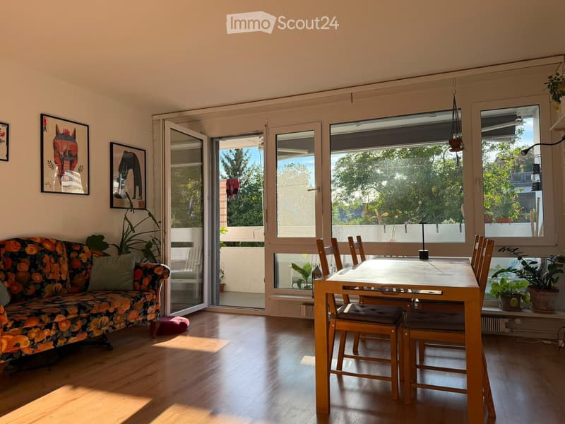 Bright living and dining area with wooden flooring, large windows, and a balcony. Features a floral-patterned sofa, dining table with chairs, and potted plants.