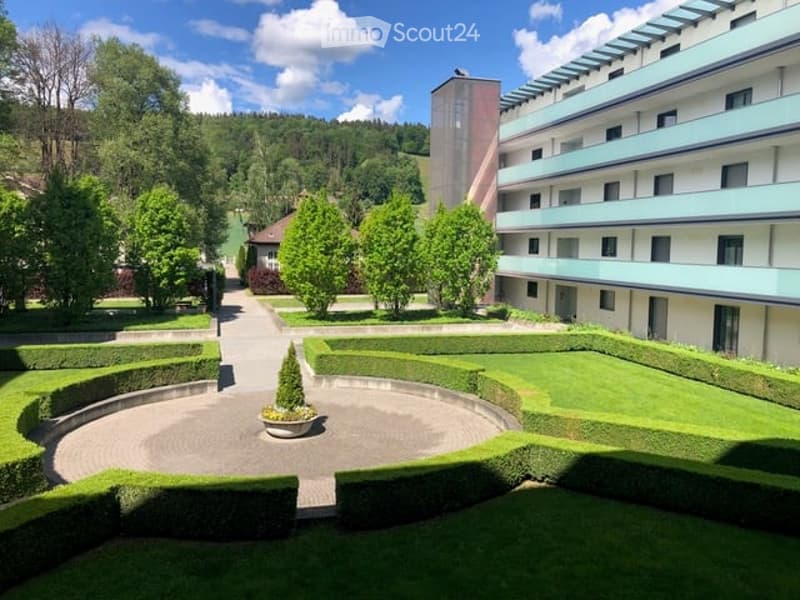 Well-maintained garden area with neatly trimmed hedges, a central circular pathway, and a small potted tree in the center. The garden is surrounded by a modern multi-story building with balconies.