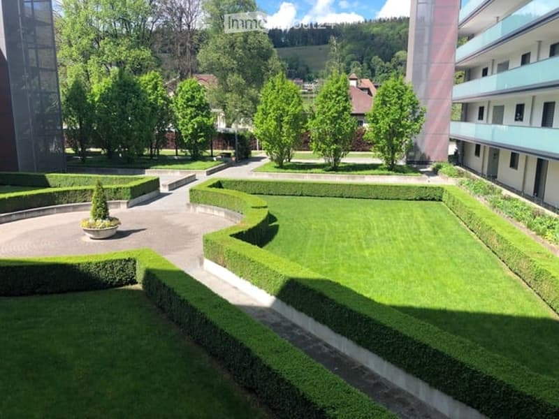 Well-maintained garden area with neatly trimmed hedges, a small tree in a pot, and a curved pathway. The area is surrounded by modern buildings and lush greenery.