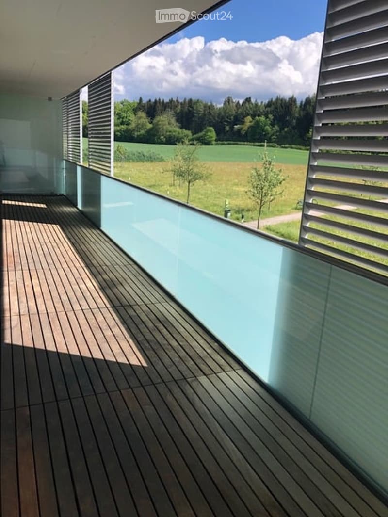 Modern balcony with wooden decking, large glass railing, and scenic countryside view.