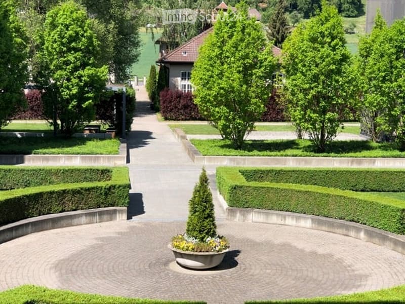 Well-maintained garden with neatly trimmed hedges, a central circular pathway, a small fountain, and surrounding trees and shrubs.