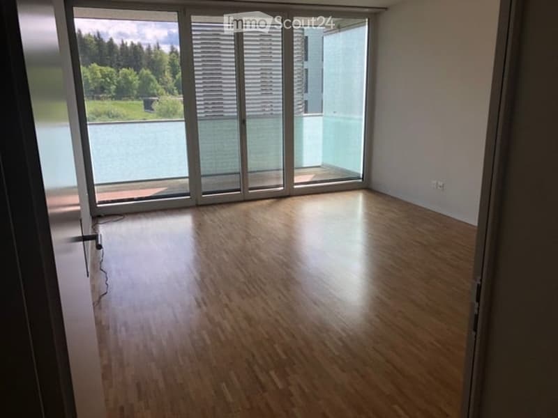 Empty room with wooden flooring and large glass sliding doors leading to a balcony with a scenic view of greenery and water.