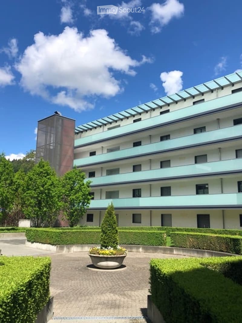 Modern multi-story residential building with light blue balconies and white walls, surrounded by neatly trimmed hedges and a central planter.