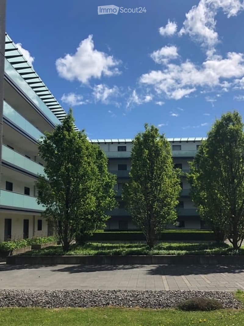 Modern multi-story building with a clean, contemporary design, surrounded by well-maintained greenery and trees. The building features large windows and a flat roof.