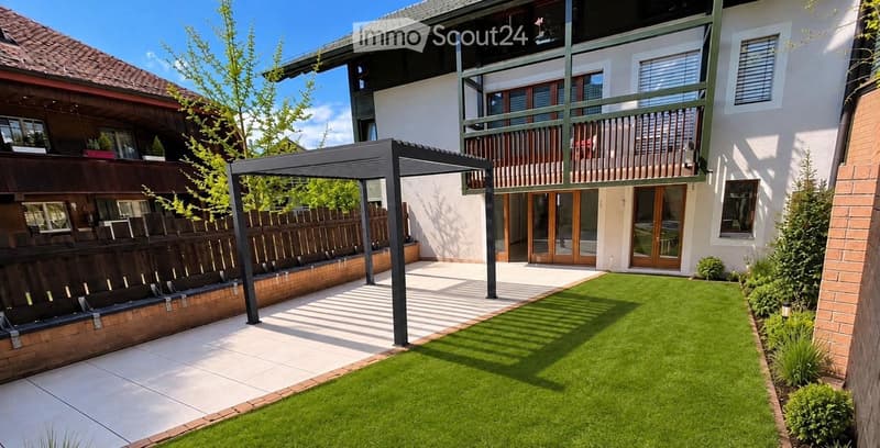 Well-maintained garden with artificial grass, a patio area, a wooden fence, and a modern pergola structure. The house features large windows and a balcony with railings.