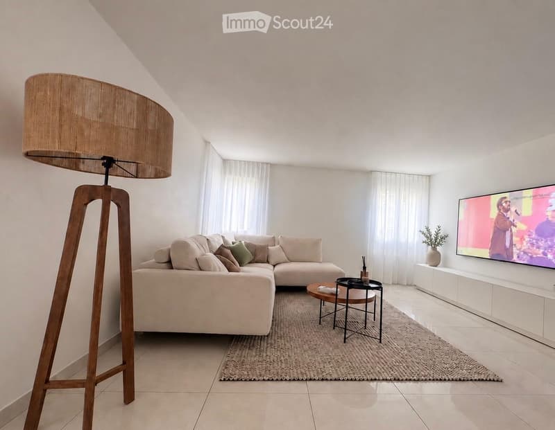 Modern living room with a large sectional sofa, coffee table, floor lamp, television, and side table with a plant. The room features large windows with white curtains and a neutral color palette.
