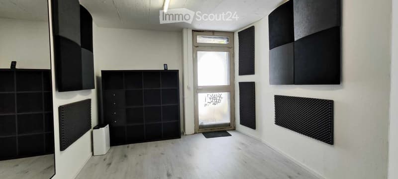 Empty room with white walls, wooden floor, black acoustic panels, and a large window with a door leading outside.