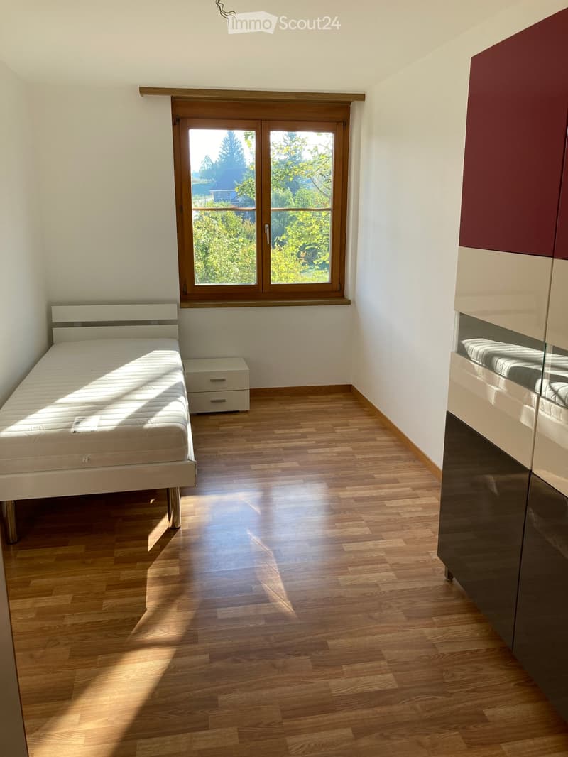 Bright bedroom with wooden flooring, a white bed, a nightstand, and a large window with wooden frame offering a view of greenery.