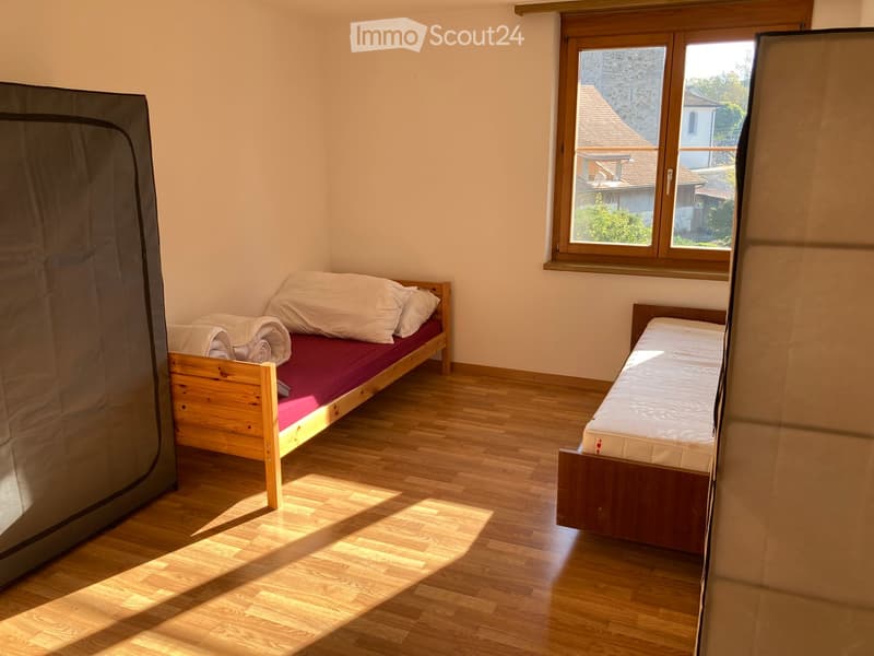 Cozy bedroom with wooden floors, two single beds, one with a red mattress and the other with a white mattress, and a window offering a view of the outside.