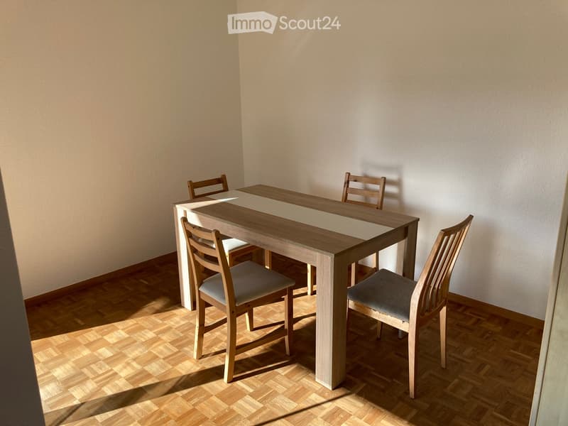 Dining room with a wooden dining table and four matching chairs, placed on a wooden floor against a plain wall.