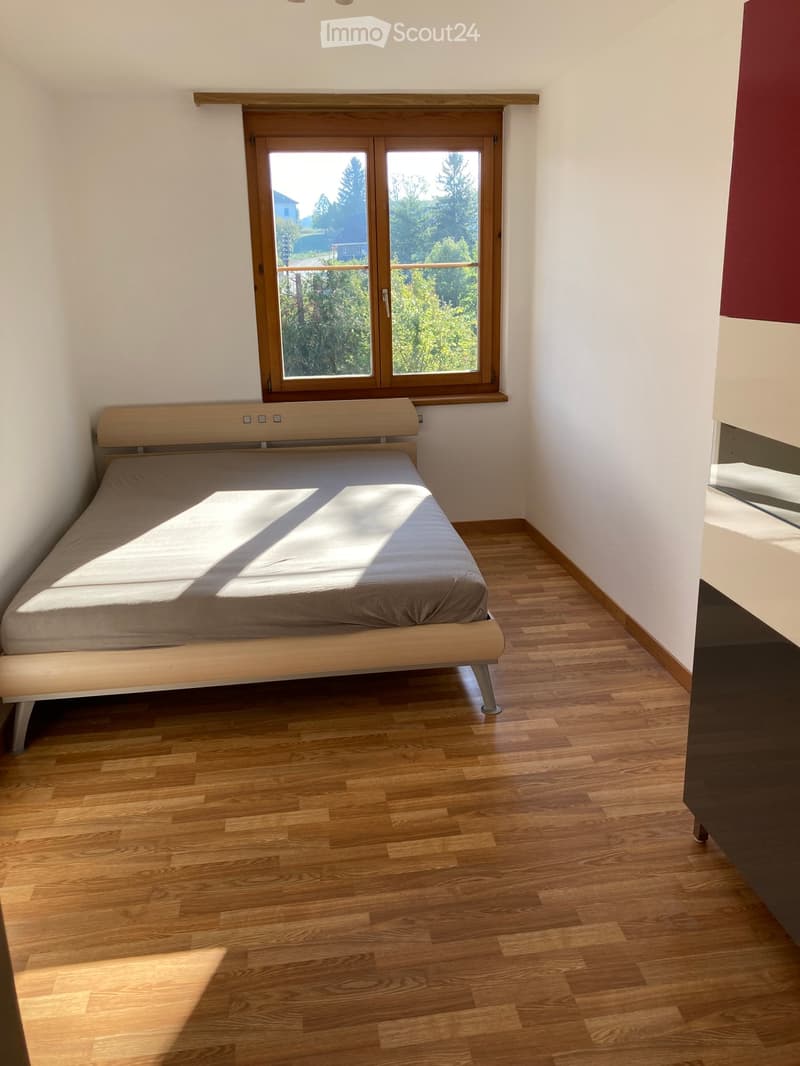 Minimalist bedroom with a single bed, wooden flooring, and large window with wooden frame offering a view of greenery.