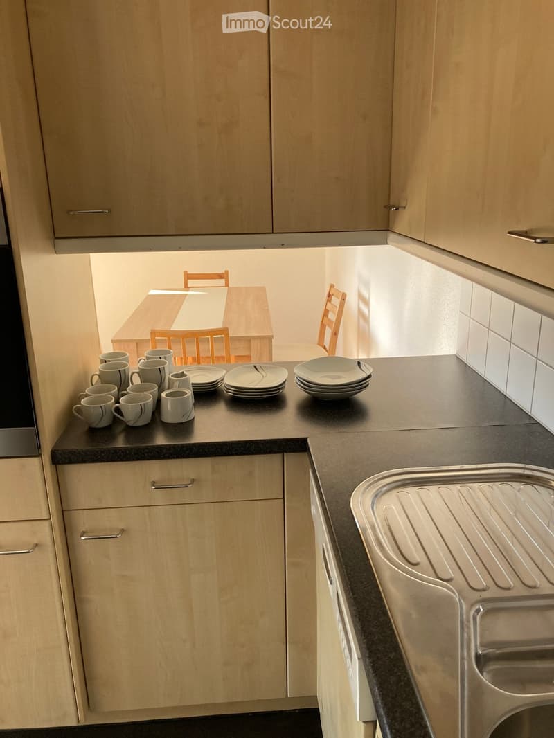 Modern kitchen with light wood cabinets, dark countertops, and a small dining area visible in the background.