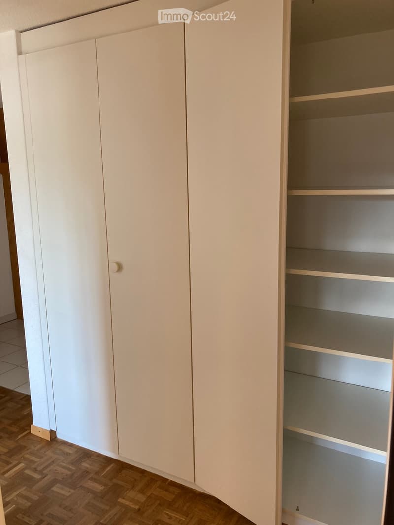 Empty room with white built-in cabinets and wooden flooring. One cabinet door is open, revealing empty shelves.