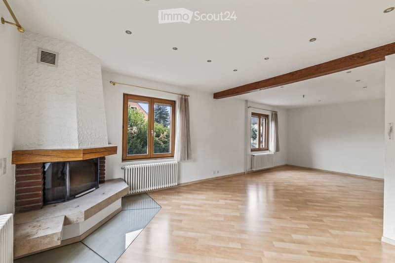 Empty living room with wooden flooring, white walls, exposed wooden beams, fireplace, large windows with curtains, and radiators.