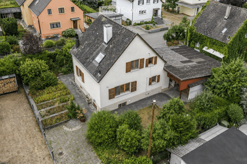 Aerial view of a two-story house with a gray roof, white walls, and brown shutters. The house features a small garden with various plants and a paved pathway leading to the entrance. Surrounding the house are other residential buildings and greenery.