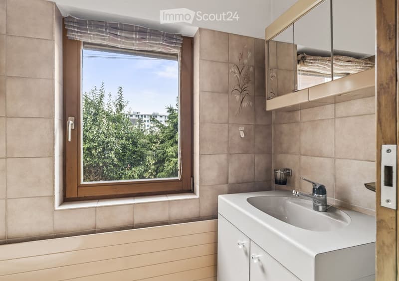 Bathroom with beige tiled walls, a white sink with a modern faucet, a mirror with a wooden frame, and a window with a view of greenery outside.