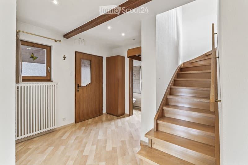 Bright hallway with wooden flooring, white walls, and a staircase leading upwards. Features include a window with decorative glass, a radiator, a wooden door, and a small wooden cabinet.