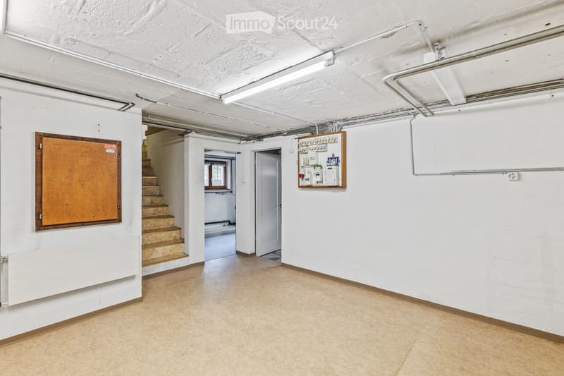 Empty basement room with white walls, tiled floor, exposed pipes, and a staircase leading upwards. A small window and a door are visible.