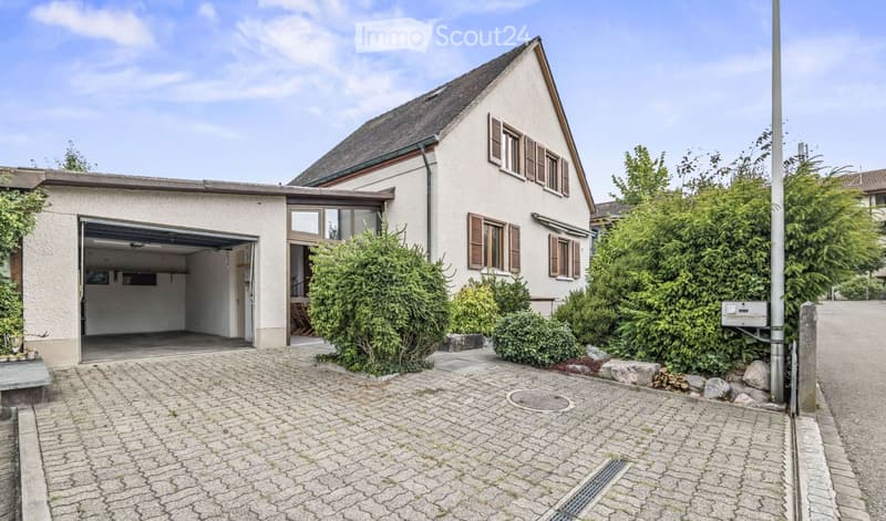 Two-story house with a garage, surrounded by greenery and a paved driveway.