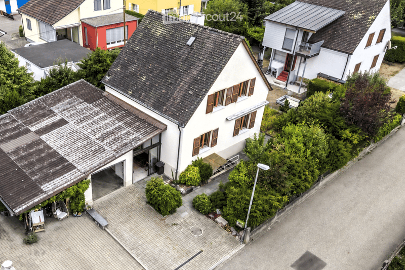 Aerial view of a two-story house with a tiled roof, white exterior, and a visible garage. The house is surrounded by greenery and neighboring houses.