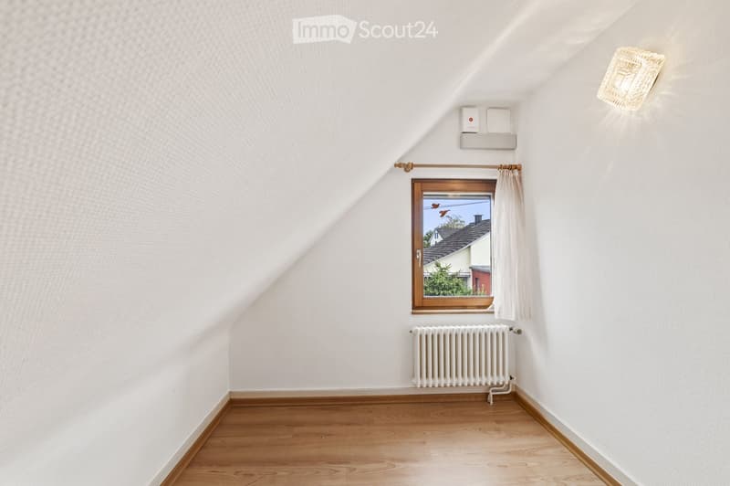 Empty attic room with wooden flooring, white walls, a window with white curtains, a radiator, and a light fixture on the wall.