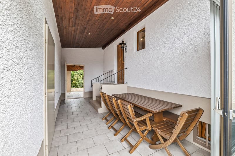 Balcony with wooden table and chairs, tiled floor, wooden ceiling, and access to a staircase leading to another level.